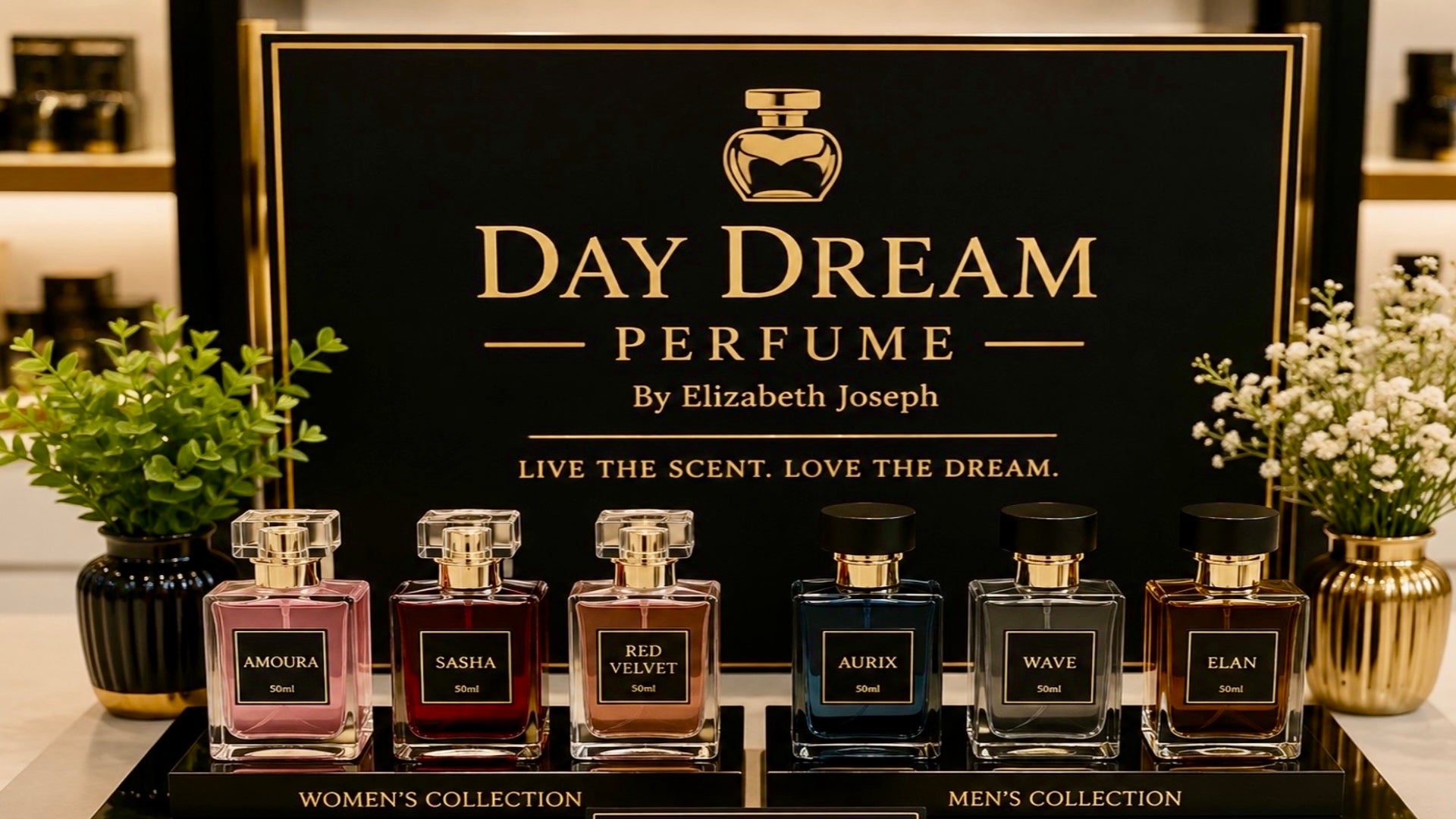 A perfume collection display with multiple glass fragrance bottles arranged in front of a large black sign reading 'DAY DREAM PERFUME By Elizabeth Joseph.' The bottles are grouped into women's and men's collections, with pink, burgundy, blue, and dark amber liquids visible inside clear rectangular bottles with gold caps.